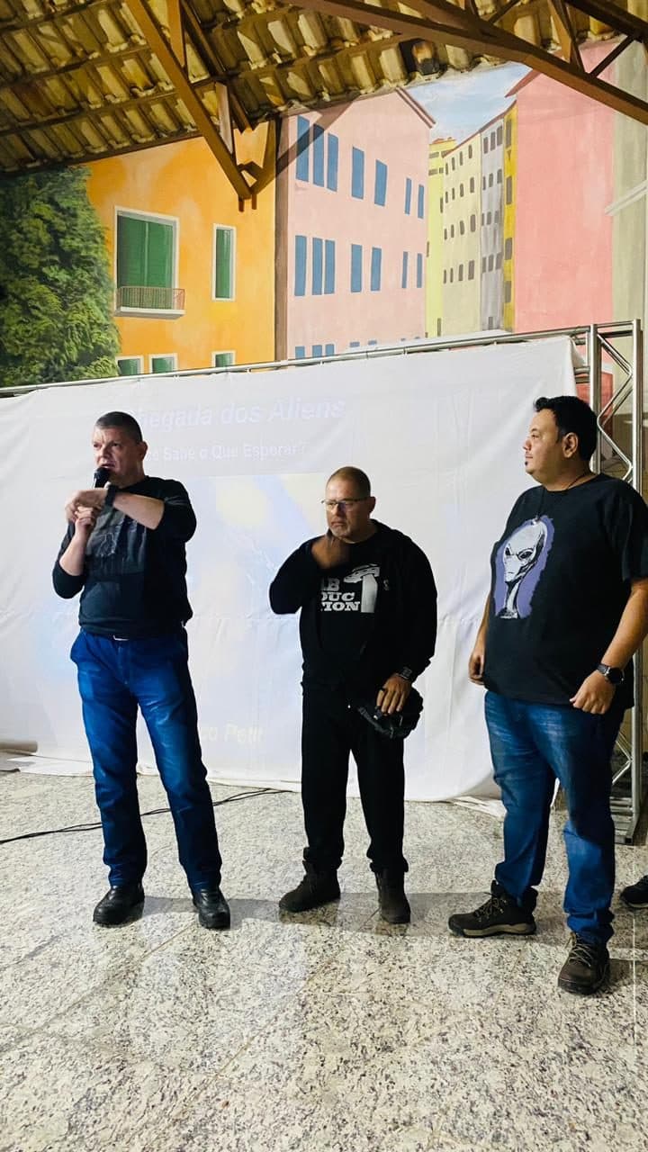 Três palestrantes de pé diante da tela, um deles ao microfone
