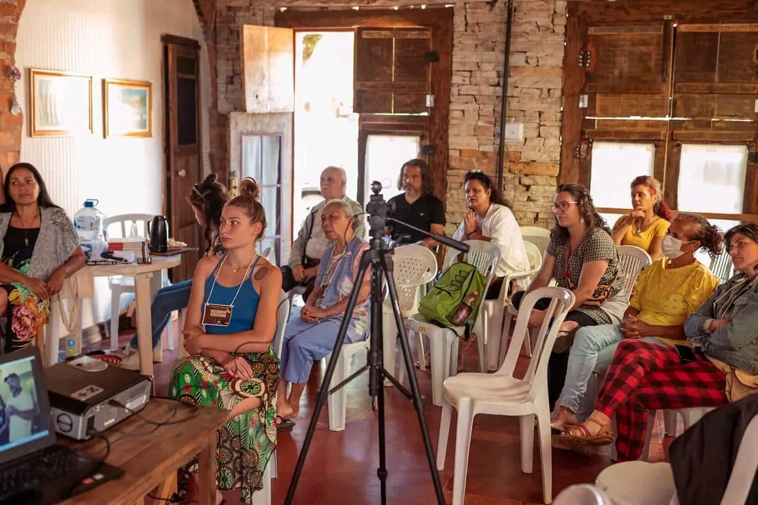 Participantes sentados em roda numa sala de paredes de pedra, com uma câmera no tripé