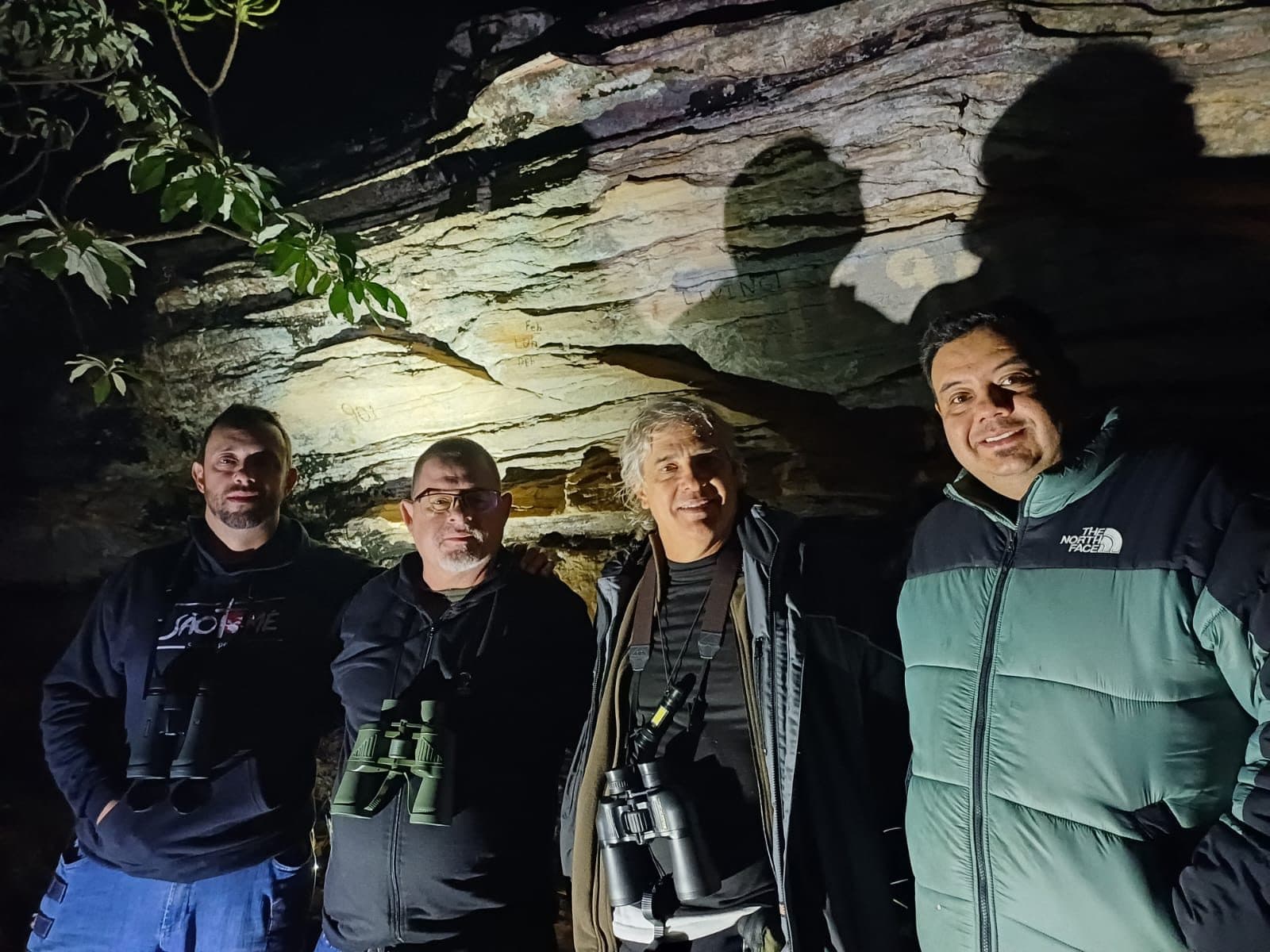 Quatro participantes com binóculos diante da pedra, iluminados por lanterna