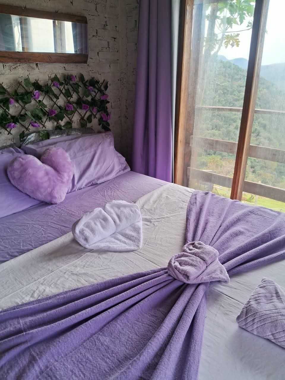 Cama de casal com manta lilás e almofada de coração, ao lado da janela aberta para a serra