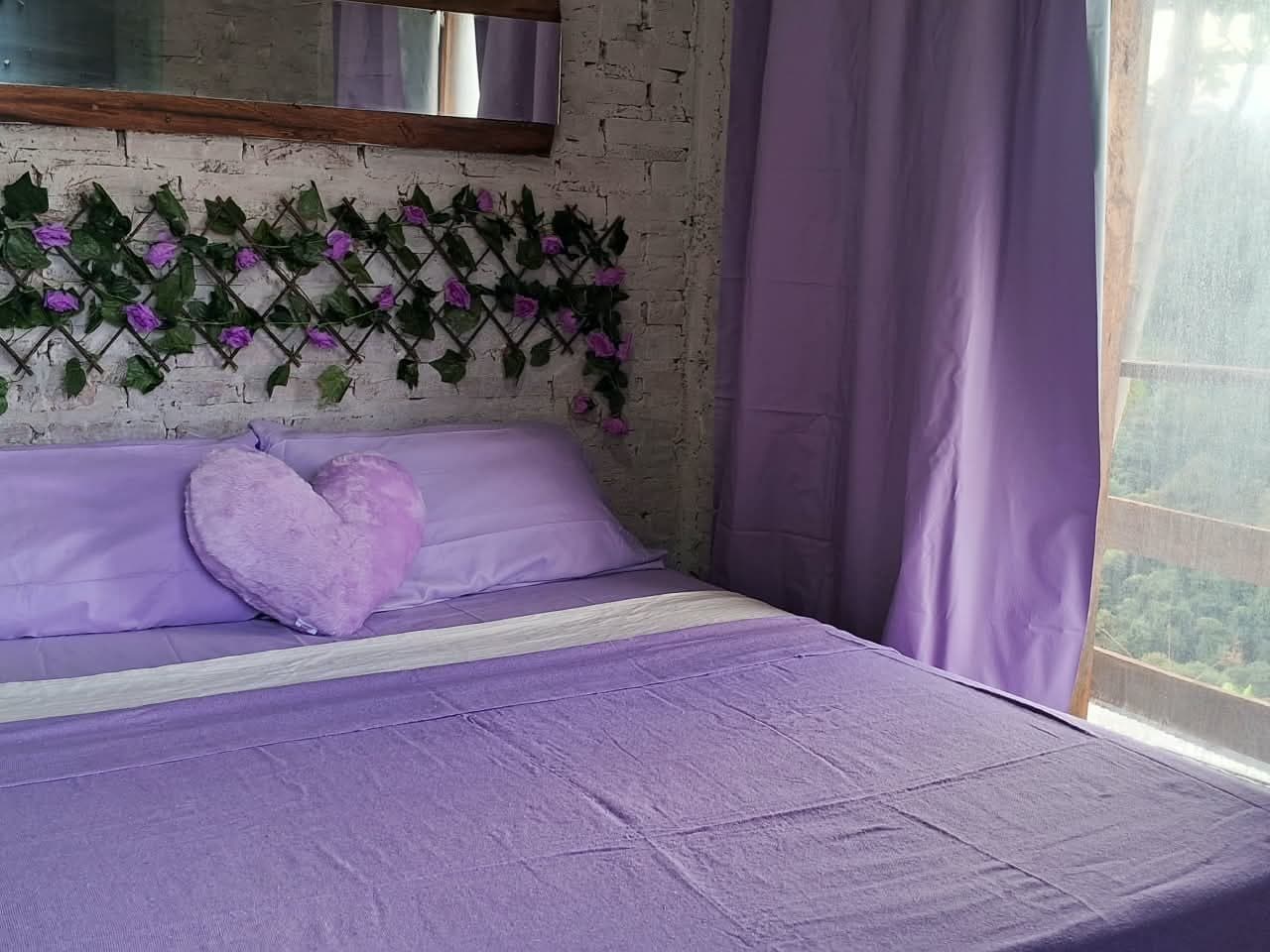 Cama com almofada de coração lilás e cortina roxa ao lado da janela