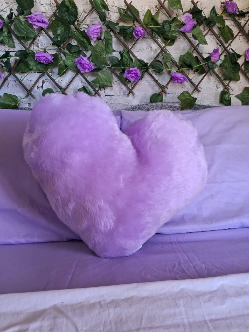 Almofada de coração lilás sobre a cama, diante da parede de flores roxas